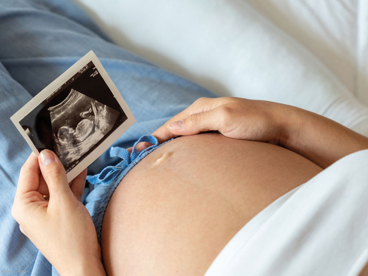 Eine schwangere Person hält im Liegen ein Ultraschallbild eines Fötus auf ihrem nackten Bauch und trägt eine blaue Hose, was auf Vorfreude und Sorgfalt während der Schwangerschaft hindeutet.