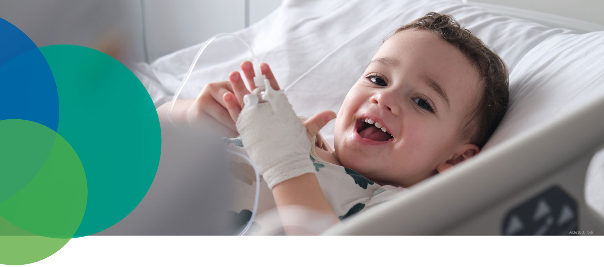 Ein kleines Kind liegt in einem Krankenhausbett, lächelt und blickt in die Kamera, seine Hand ist bandagiert und an eine Infusion angeschlossen. Der Hintergrund besteht aus bunten, sich überschneidenden Kreisen auf der linken Seite.