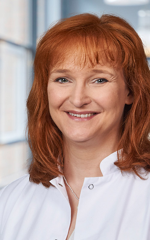 Eine lächelnde Frau mit schulterlangem rotem Haar, die einen weißen Arztkittel trägt, steht in einem gut beleuchteten Raum mit Fenstern im Hintergrund.
