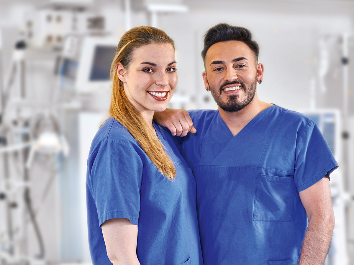 Zwei Fachkräfte des Gesundheitswesens in blauen Kitteln lächeln und stehen dicht beieinander in einem Krankenhaus oder einer medizinischen Einrichtung, wobei die medizinischen Geräte im unscharfen Hintergrund sichtbar sind.