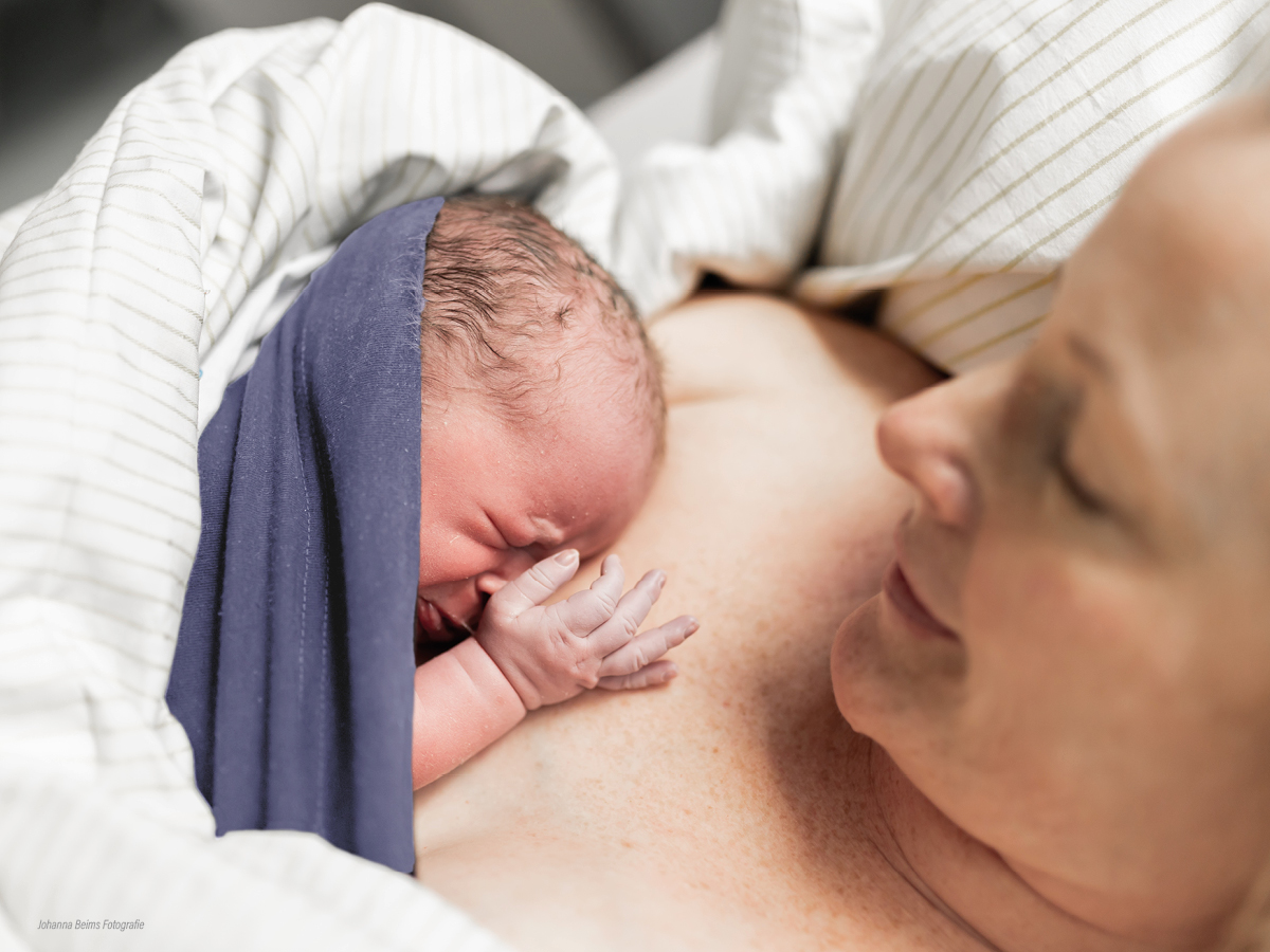 Ein neugeborenes Baby, eingewickelt in ein dunkelblaues Tuch, ruht auf der nackten Brust einer lächelnden Frau, beide unter einer gestreiften Decke, in einem zärtlichen Moment unmittelbar nach der Geburt.