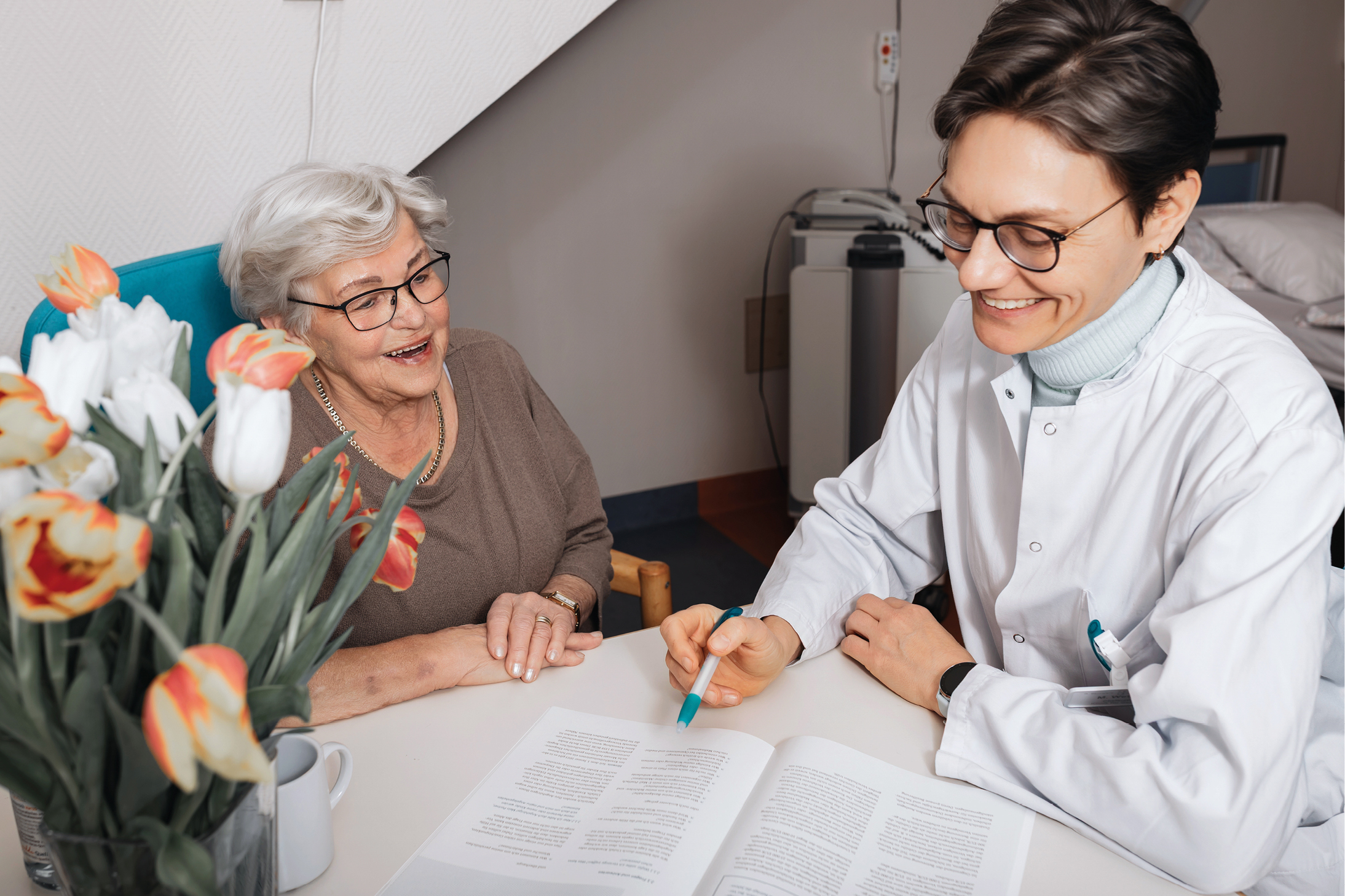 Eine ältere Frau sitzt an einem Tisch mit einer lächelnden medizinischen Fachkraft, die einen weißen Kittel trägt und einen Stift hält, während sie Dokumente bespricht. Eine Vase mit Tulpen steht auf dem Tisch.