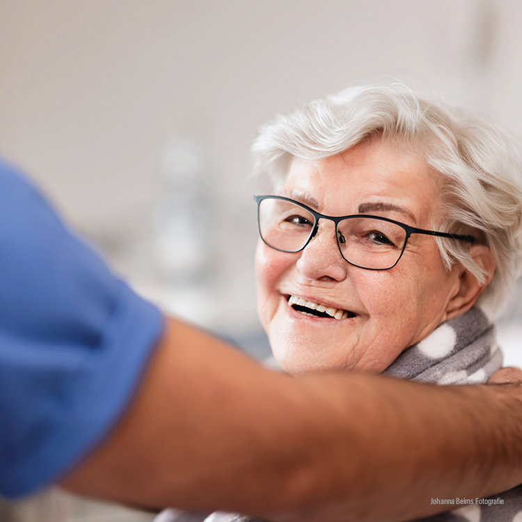 Eine ältere Frau mit kurzen grauen Haaren und Brille lächelt warm in die Kamera. Sie ist in eine kuschelige Decke eingewickelt, und der Arm einer Person, möglicherweise einer Pflegekraft, ruht sanft auf ihrer Schulter.