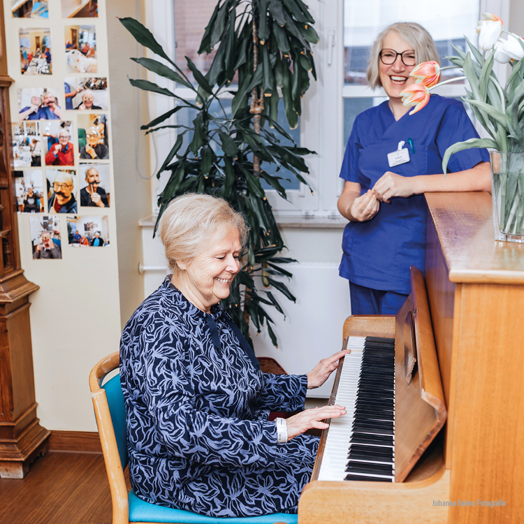 Eine ältere Frau spielt lächelnd auf einem Klavier, während eine Krankenschwester in blauem Kittel daneben steht und ebenfalls lächelt. Sie befinden sich in einem hellen Raum mit Fotos an der Wand und einer Vase mit Blumen auf dem Klavier.