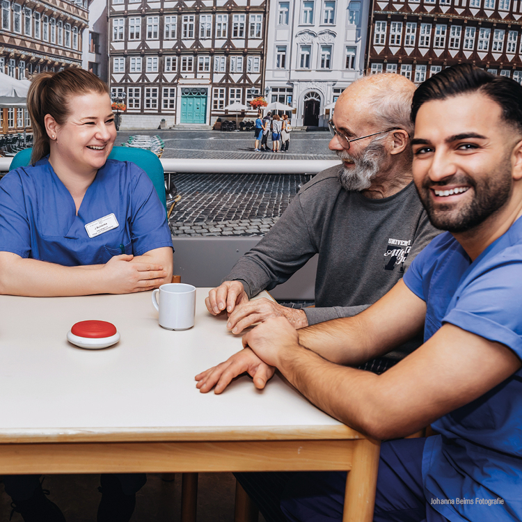 Zwei Mitarbeiter des Gesundheitswesens sitzen mit einem älteren Mann an einem Tisch, lächeln und unterhalten sich. Auf dem Tisch befinden sich ein weißer Becher und ein roter Knopf. Im Hintergrund sind traditionelle Fachwerkhäuser zu sehen.