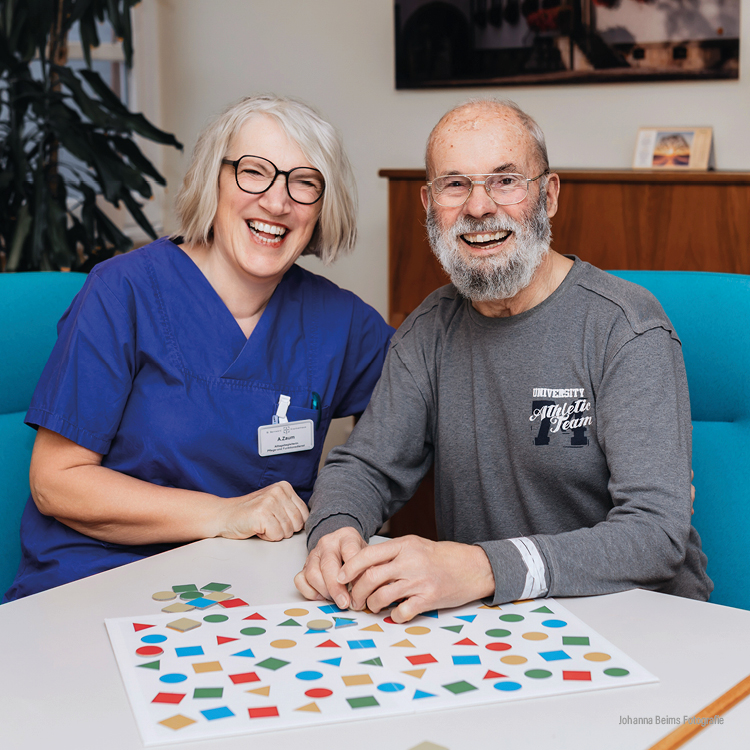 Eine lächelnde Krankenschwester im blauen Kittel sitzt neben einem älteren Mann mit Bart. Sie sitzen an einem Tisch mit einem farbenfrohen Puzzle mit geometrischen Formen und wirken glücklich und in eine gemeinsame Aktivität vertieft.
