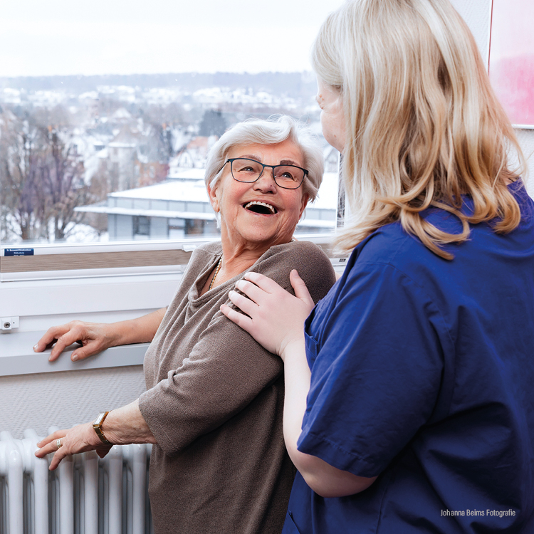 Eine ältere Frau lächelt und lacht, während sie an einem Fenster steht. Eine Pflegekraft in blauem Kittel hält sanft ihren Arm und stützt sie. Im Hintergrund sind verschneite Gebäude und Bäume außerhalb des Fensters zu sehen.