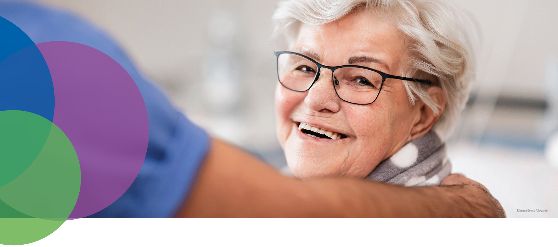 Eine ältere Frau mit kurzen grauen Haaren und Brille lächelt warm und schaut in die Kamera. Eine Person in einem blauen Hemd legt ihr sanft eine Hand auf die Schulter. Bunte, sich überlappende Kreise zieren die linke Seite des Bildes.