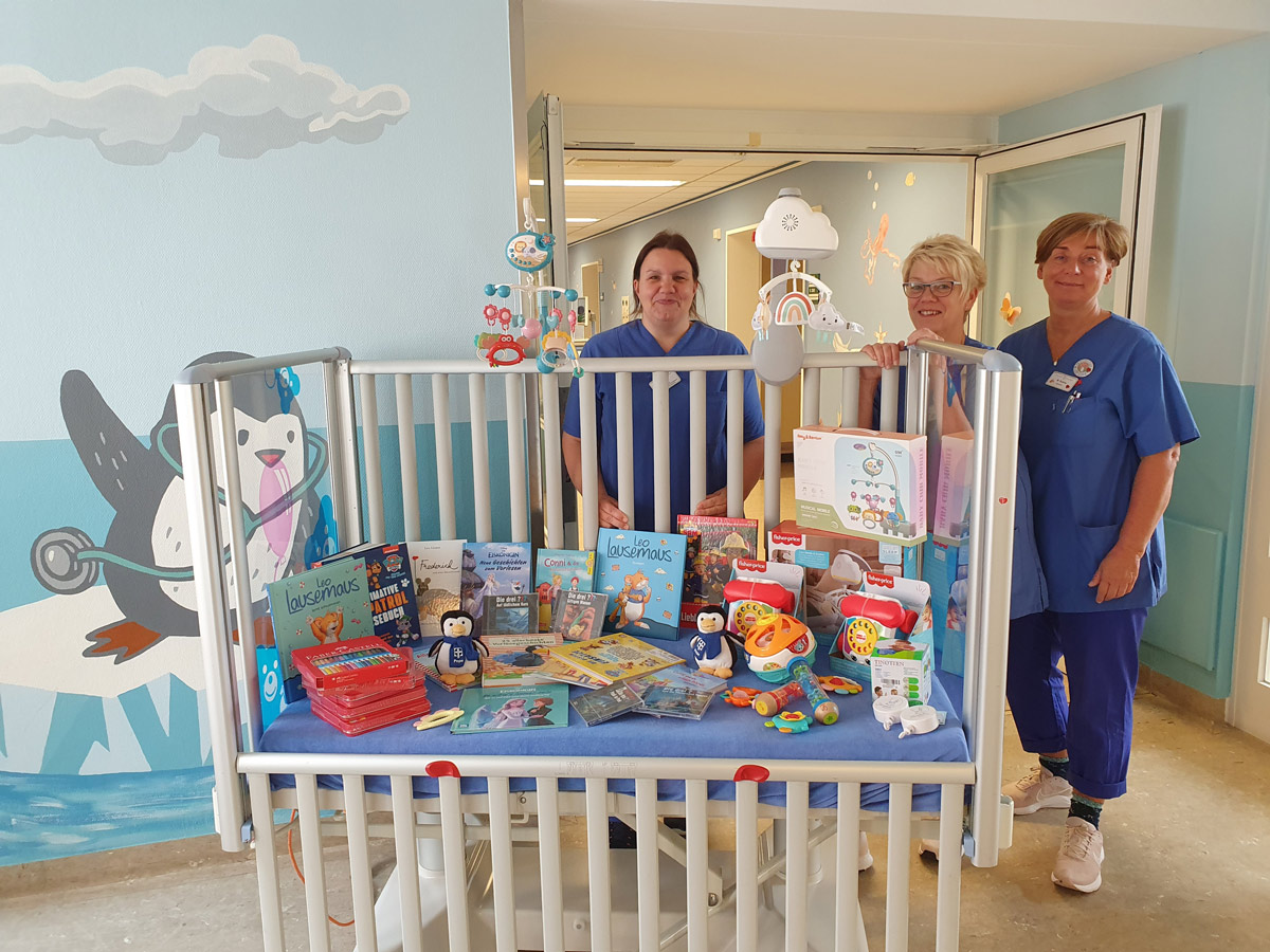 Drei Mitarbeiter des Gesundheitswesens stehen hinter einem Krankenhausbett voller Bücher, Spielzeug und Spiele in einem hell dekorierten Raum mit Pinguin- und arktischen Tierwandbildern.