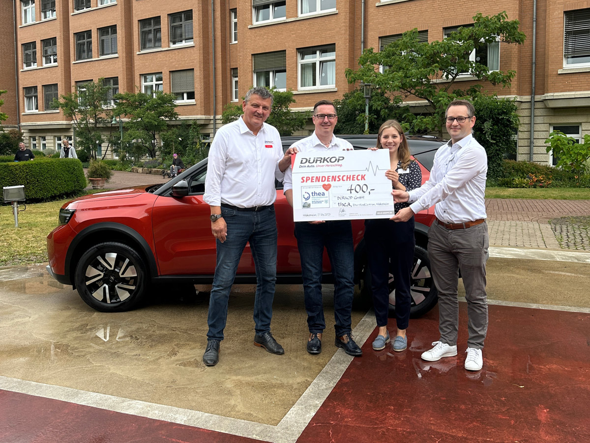 Vier Personen stehen vor einem roten Auto und halten einen großen Spendenscheck in der Hand. Sie lächeln und scheinen vor einem Backsteingebäude zu stehen, das im Hintergrund begrünt ist.