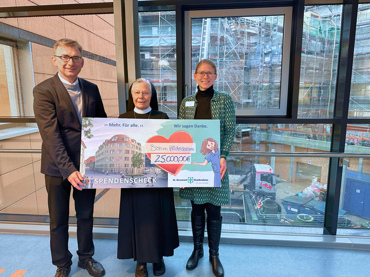 Drei Personen stehen in einem Innenraum und halten einen großen Spendenscheck über 250.000 Euro für das Bistum Hildesheim in der Hand. Hinter ihnen ist ein großes Fenster mit Blick auf eine Baustelle mit Gerüsten und Baumaterialien.
