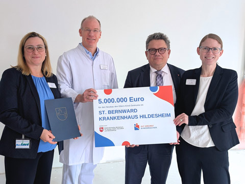 Vier Personen stehen zusammen und halten lächelnd einen großen Scheck über 5.000.000 Euro und eine Urkunde für das St. Bernward Krankenhaus Hildesheim in der Hand. Eine Person trägt einen weißen Arztkittel, die anderen tragen Business-Kleidung.