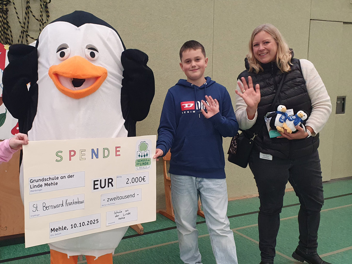 Eine Person im Pinguinkostüm, ein lächelnder Junge und eine Frau mit Plüschpinguinen stehen in der Halle, winken und halten einen großen Spendenscheck über 2.000 Euro für das St. Bernward Krankenhaus.