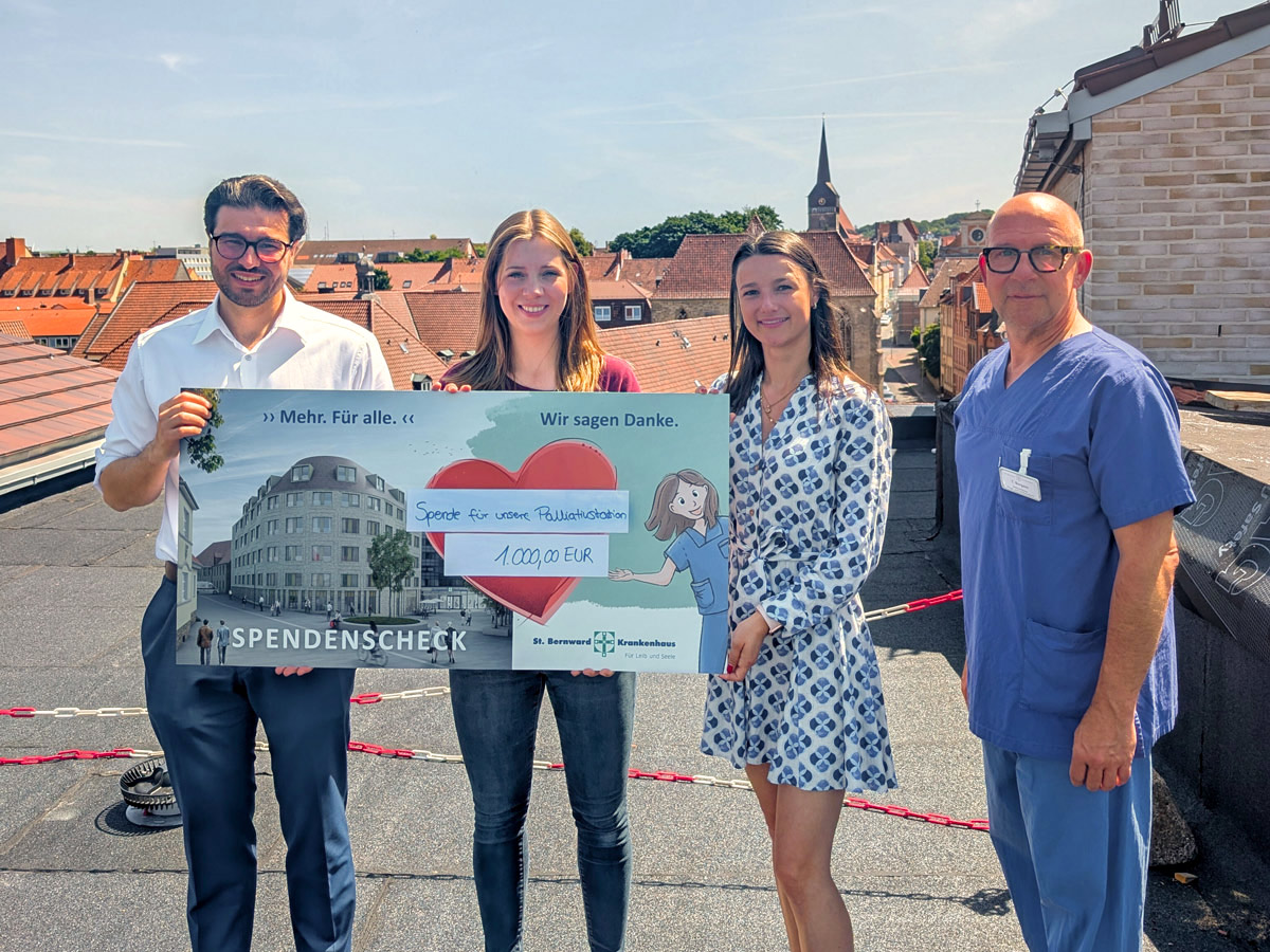 Vier Personen stehen auf einem Dach und halten einen großen Spendenscheck über 1.000 Euro in der Hand, im Hintergrund eine Stadtlandschaft mit Kirchturm. Der Scheck enthält Zeichnungen und den Dank für die Unterstützung der Palliativarbeit.
