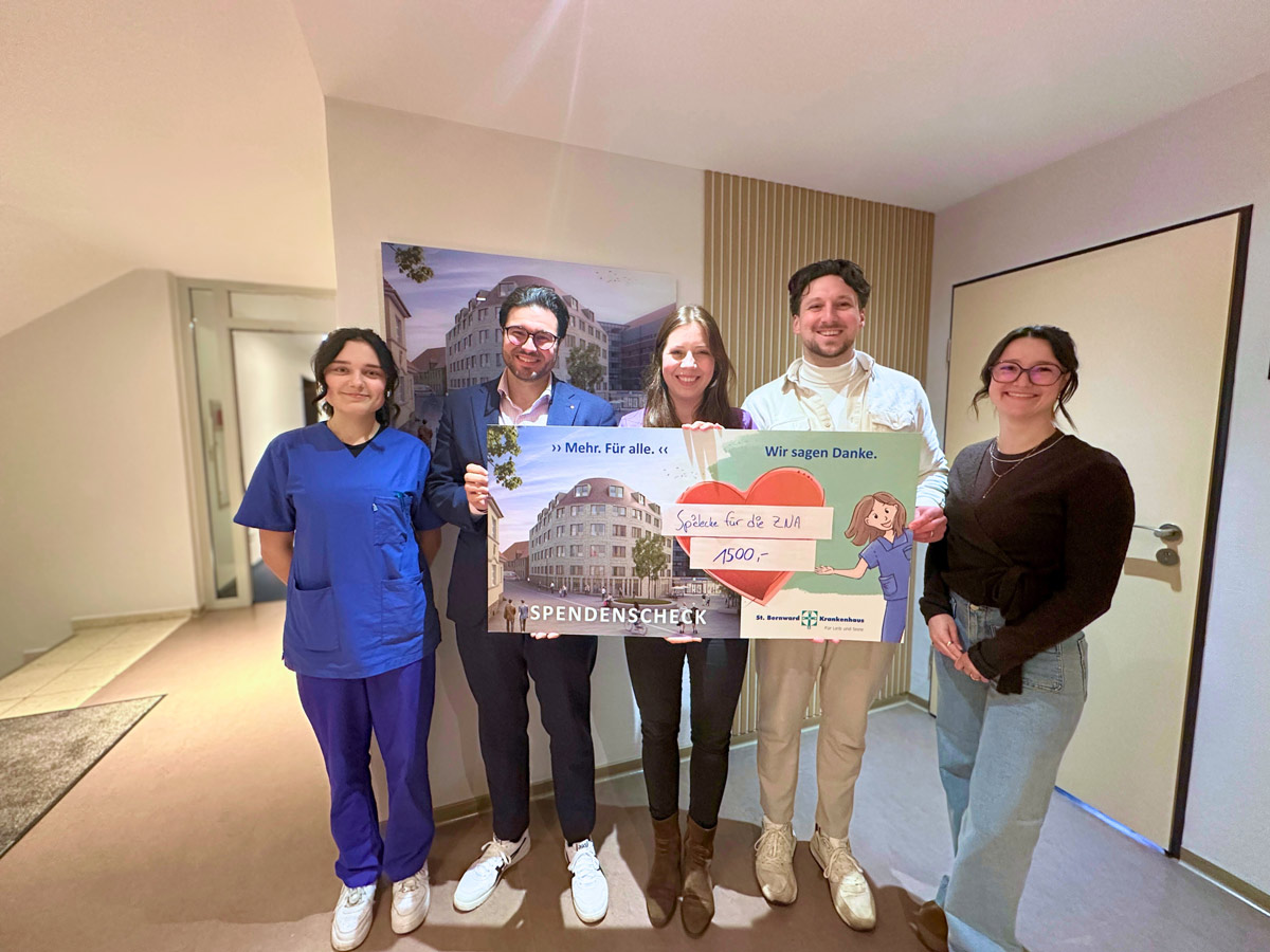 Fünf Personen stehen drinnen und lächeln in die Kamera; zwei Personen in der Mitte halten einen großen Spendenscheck. Eine Person trägt einen medizinischen Kittel, die anderen tragen Freizeit- oder Business-Casual-Kleidung.