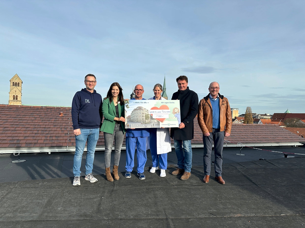 Sechs Personen stehen auf einem Dach und halten einen großen feierlichen Scheck. Einige tragen medizinische Kittel, die anderen Freizeit- oder Geschäftskleidung. Im Hintergrund sind bei klarem Himmel Kirchtürme und Gebäude zu sehen.