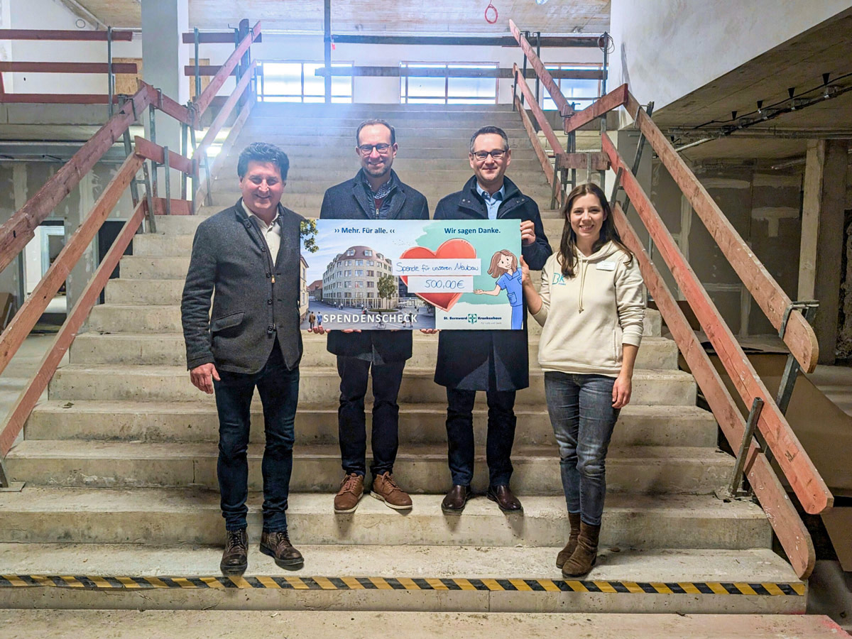 Vier Personen stehen auf einer Betontreppe in einem unfertigen Gebäude, lächeln und halten einen großen Scheck über 500 Euro in der Hand, auf dem ein Spendenschild und ein Foto eines Gebäudes aufgedruckt sind.