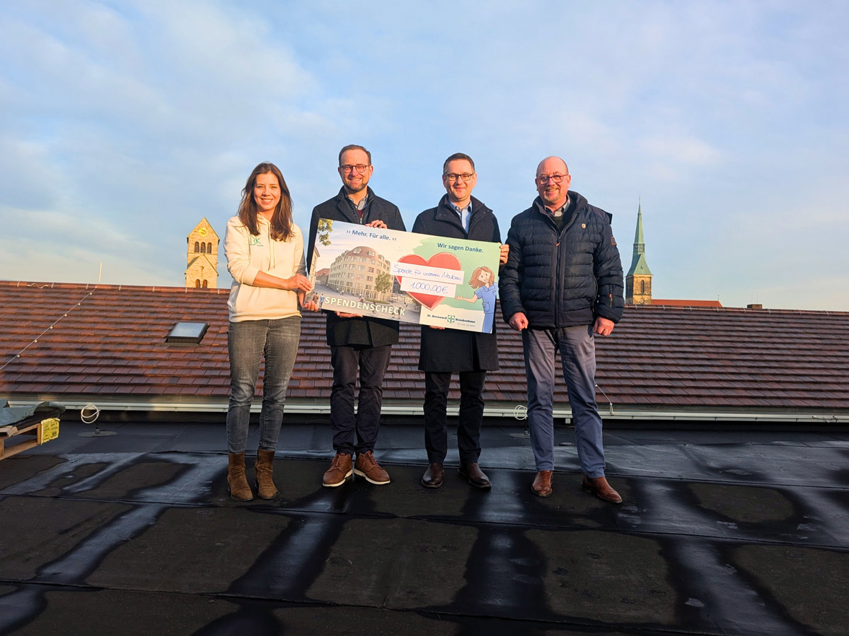 Vier Personen stehen auf einem Dach und halten einen großen feierlichen Scheck. Im Hintergrund sind zwei Kirchtürme bei teilweise bewölktem Himmel zu sehen. Die Gruppe lächelt und ist in Winterkleidung gekleidet.