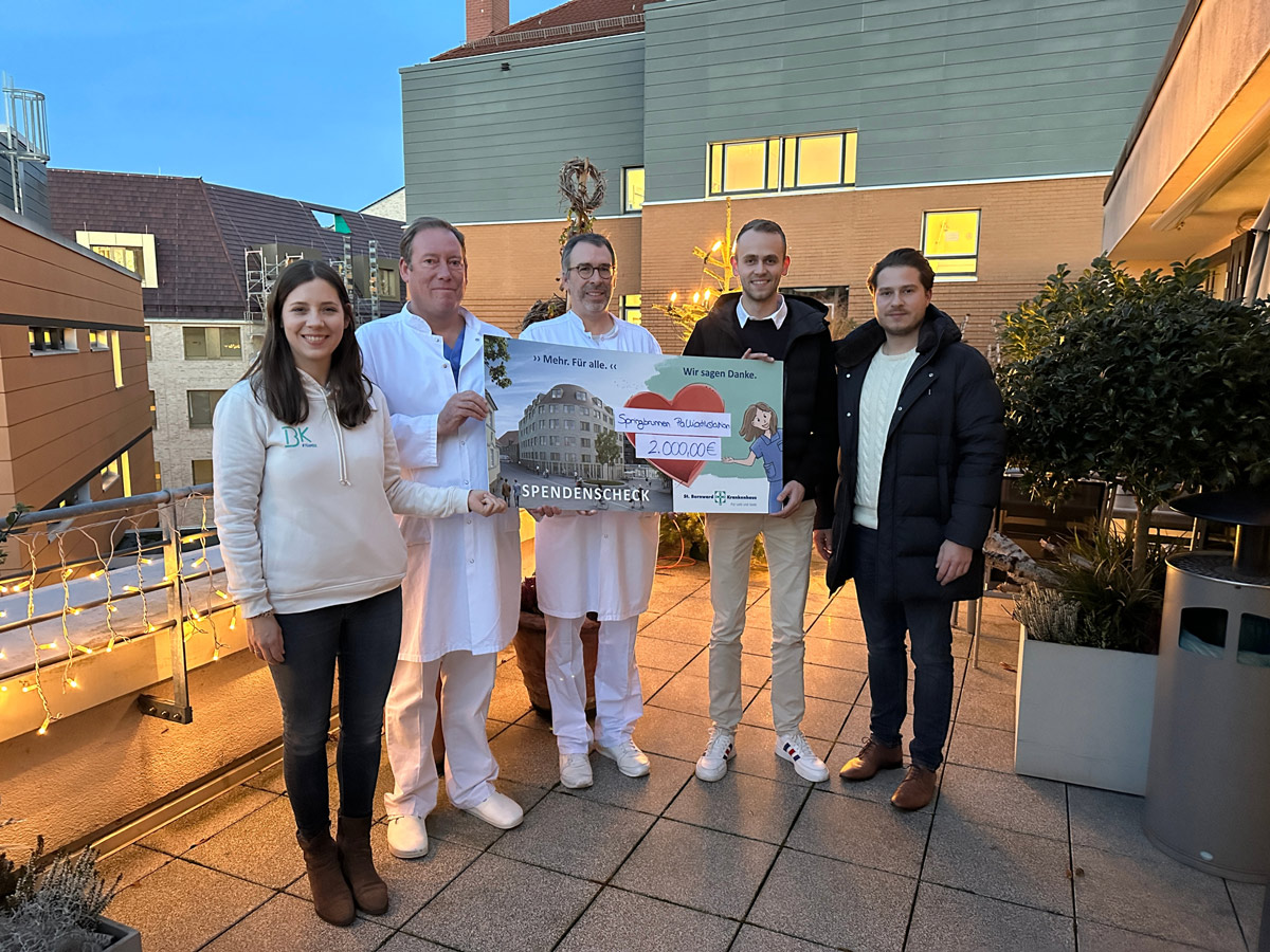 Fünf Personen stehen auf einer Dachterrasse, zwei davon in weißen Arztkitteln, und halten einen großen Scheck über 2.000 Euro in der Hand. Im Hintergrund sind Lichterketten und Gebäude zu sehen.