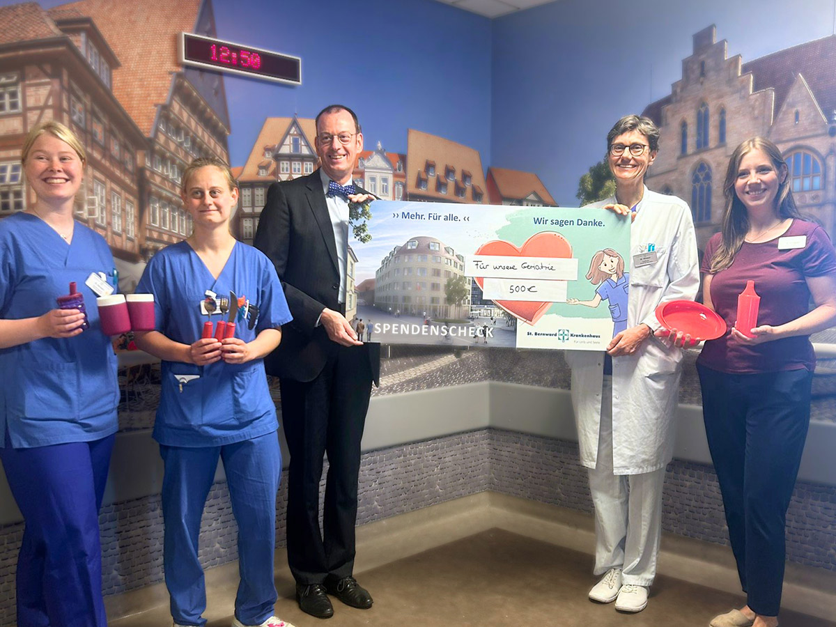 Fünf Personen, darunter medizinisches Personal, posieren in einem Krankenhauszimmer und halten einen großen Spendenscheck über 500 Euro in den Händen. Einige halten Tassen und einen Teller in den Händen. Im Hintergrund ist ein Wandgemälde mit Gebäuden zu sehen.