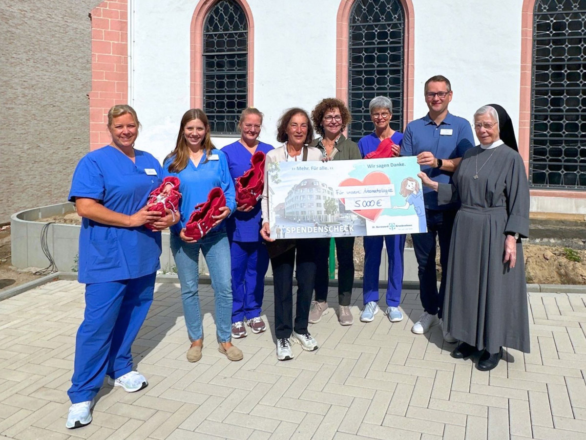 Eine Gruppe von Menschen, darunter medizinisches Personal und eine Nonne, steht draußen, hält einen großen Spendenscheck über 500 Euro sowie Körbe voller roter Gegenstände in den Händen und lächelt in die Kamera.