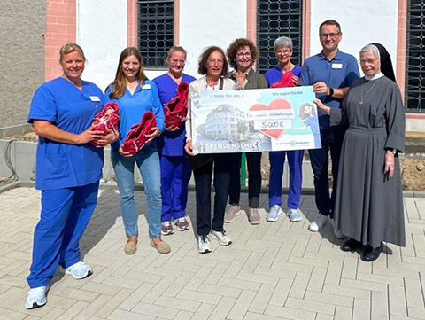 Eine Gruppe von acht Personen, darunter eine Nonne, hält vor einem Kirchengebäude einen großen Spendenscheck über 5.000 Euro in den Händen.