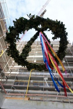 Ein großer, mit grünen Blättern und bunten Bändern geschmückter Kranz hängt hoch über einer Baustelle, umgeben von Baugerüsten und teilweise fertiggestellten Gebäuden.