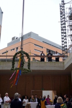 Eine Gruppe von Menschen steht auf einer Baustelle und beobachtet, wie ein Kranz mit bunten Bändern von einem Kran über ihnen herabhängt. Einige Personen befinden sich auf einer oberen Ebene, und rechts ist ein Baugerüst zu sehen.
