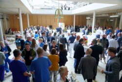 Eine große Gruppe von Menschen, die sich in einem geräumigen, unfertigen Gebäude mit natürlichem Licht von oben um Tische mit Getränken und Snacks versammelt hat.