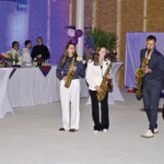 Eine Gruppe von vier Musikern, drei Frauen und ein Mann, spielen drinnen Saxophone, während ein Junge auf einer Cajón-Trommel sitzt. Hinter ihnen stehen Cocktailtische, Menschen, die sich unterhalten, und Veranstaltungsdekorationen.