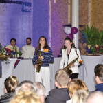 Eine Gruppe von Musikern mit Saxophonen steht bei einer Indoor-Veranstaltung vor einem Publikum. Drei Frauen und ein Mann lächeln auf der Bühne, während die Gäste ihnen gegenüber sitzen. Im Hintergrund sind eine Bar und dekorierte Tische zu sehen.
