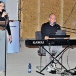 Eine Frau singt in ein Mikrofon, während sie neben einem Mann steht, der ein Kawai ES7 Keyboard spielt. Sie spielen in einem Innenraum, in dem Kabel, eine Wasserflasche und Pflanzen im Hintergrund zu sehen sind.