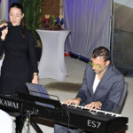 Eine Frau in einem schwarzen Kleid singt in ein Mikrofon, während ein Mann im Anzug und mit gelber Brille ein Kawai ES7 Keyboard spielt. Sie treten in einem Innenraum neben Stehtischen und Pflanzen auf, was wie eine gesellschaftliche Veranstaltung aussieht.