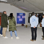 Eine Gruppe von Menschen steht in einem Betonraum, einige vor einer blauen Schautafel mit Bildern und Informationen. Zwei Personen tragen hellblaue Kapuzenpullover mit BK auf dem Rücken. Die Atmosphäre wirkt informell und aufmerksam.