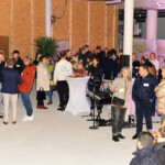 Eine Gruppe von Menschen ist in einem Raum bei einer gesellschaftlichen Veranstaltung oder einem Networking-Event versammelt. Einige stehen und unterhalten sich an Cocktailtischen, während andere sitzen. Die Umgebung wirkt unfertig, mit freiliegenden Wänden und sichtbaren Baumaterialien.