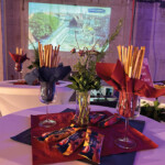 Ein runder Tisch mit Brotstangen in Weingläsern, eingewickelt in rote und blaue Servietten, und eine Vase mit Blumen. Auf dem Tisch stehen Snackpakete. Im Hintergrund ist ein projiziertes Bild einer Stadtlandschaft zu sehen.