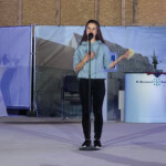 Eine Frau in einem hellblauen Kapuzenpulli steht auf einer Bühne und spricht in ein Mikrofon. Hinter ihr stehen ein gedeckter Tisch mit Blumen, ein Podium und ein Hintergrund mit einem Gebäudebild und dem Logo des St. Bernward Krankenhauses.