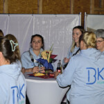Eine Gruppe von Frauen in hellblauen "BK"-Team-Kapuzenpullovern versammelt sich bei einer Indoor-Veranstaltung um einen hohen runden Tisch mit Snacks und Getränken. Einige Frauen unterhalten sich und lächeln, und die Umgebung wirkt ungezwungen.
