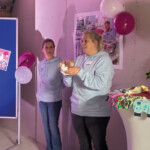 Zwei Frauen stehen in einem Haus in der Nähe einer blauen Schautafel mit Plakaten und Luftballons. Eine Frau hält eine Puppe und spricht, während die andere zuhört. Auf einem Tisch in der Nähe stehen Blumen, Snacks und dekorierte Stoffe.