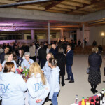 Eine große Gruppe von Menschen trifft sich bei einer Indoor-Veranstaltung in einem geräumigen, unvollendeten Raum. Einige tragen passende hellblaue Kapuzenpullover, während andere an Tischen mit Getränken und Snacks stehen oder sitzen. Die Atmosphäre ist lebhaft und ungezwungen.