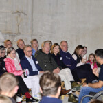 Eine Gruppe von Menschen sitzt auf Stühlen in einem Innenraum und hört aufmerksam einem Redner im Rollstuhl zu. Einige Zuhörer lächeln, andere machen Fotos oder Videos mit ihren Handys.