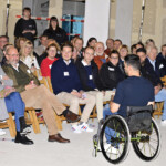 Ein Mann im Rollstuhl spricht zu einem aufmerksamen Publikum, das in Reihen von Holzstühlen bei einer Indoor-Veranstaltung sitzt. Die Zuhörer wirken engagiert, einige lächeln und interagieren mit dem Redner.