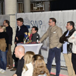 Eine Gruppe von Menschen, sowohl sitzend als auch stehend, verfolgt aufmerksam eine Veranstaltung in einem Innenraum. Einige stehen um einen hohen Tisch mit Getränken und Snacks, andere sitzen mit dem Gesicht nach vorne. Im Hintergrund sind eine Fahne und Betonpfeiler zu sehen.