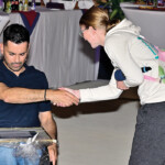 Ein Mann im Rollstuhl schüttelt einer Frau in legerer Kleidung bei einer Indoor-Veranstaltung die Hand, während im Hintergrund Tische und Flaschen zu sehen sind. Der Mann hält einen gerahmten Gegenstand auf seinem Schoß.