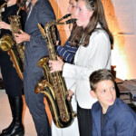 Eine Gruppe von Musikern, darunter drei Erwachsene, die Saxophon spielen, und ein Kind im Anzug, das vorne sitzt, spielen in einem Innenraum vor einem weißen Hintergrund mit warmer Beleuchtung.