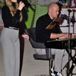 Eine Frau singt in ein Mikrofon, während ein Mann ein Keyboard spielt. Beide sind in legerer, professioneller Kleidung gekleidet. Im Hintergrund sind eine große architektonische Karte und ein Tisch mit bunten Dekorationen zu sehen.