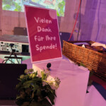 Ein Schild auf Deutsch mit der Aufschrift Vielen Dank für Ihre Spende! (Vielen Dank für Ihre Spende!) steht auf einem weißen Ständer mit Grünzeug, neben einem Korb und einem Projektor in einem dekorierten Innenraum.