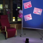 Ein roter Sessel steht neben einer blauen Pinnwand, auf der rosa und weiße Informationsplakate angebracht sind. An der Tafel ist ein rosa Luftballon befestigt. Der schummrige Raum hat Fenster im Hintergrund und einen kleinen Scheinwerfer auf dem Boden.
