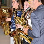 Drei Personen spielen in einem Haus Saxophon. Zwei junge Frauen stehen im Vordergrund, eine trägt einen gestreiften Pullover, und ein Mann im Anzug steht neben ihnen. Alle sind auf ihr Spiel konzentriert.